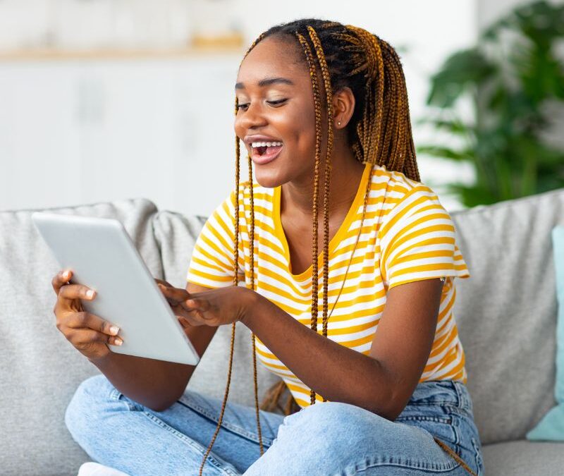Black girls sitting on a couch reading a tablet wearing a yellow shirt and blue jeans