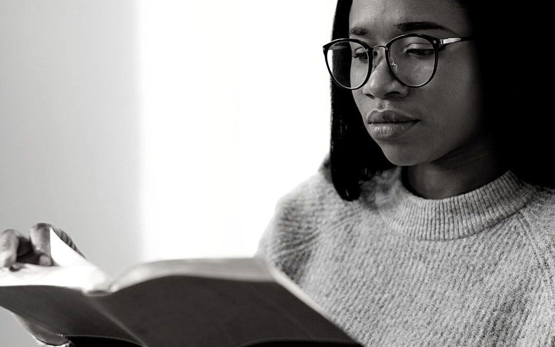 Black woman sitting on the floor reading a book. She is wearing a sweater and jeans with glasses on her face.