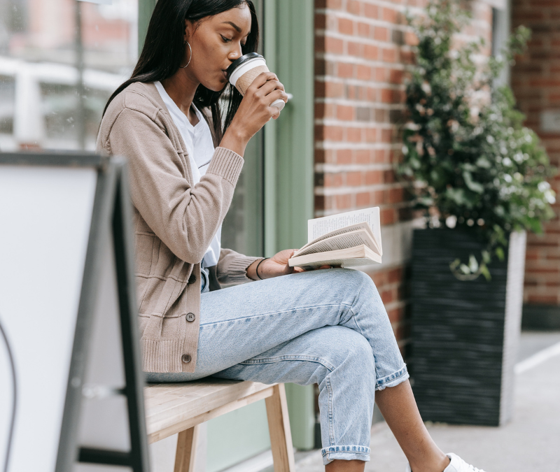 african american girl sitting outside with a cardigan and jeans reading a book with coffee